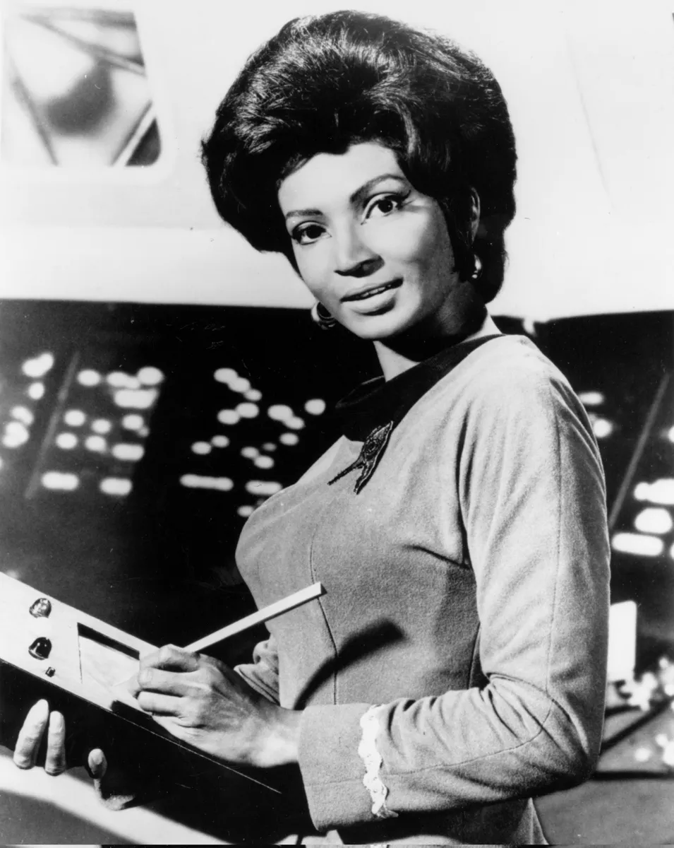 Nichelle Nichols as Uhura in a black-and-white publicity still, holding a clipboard at the communications station.