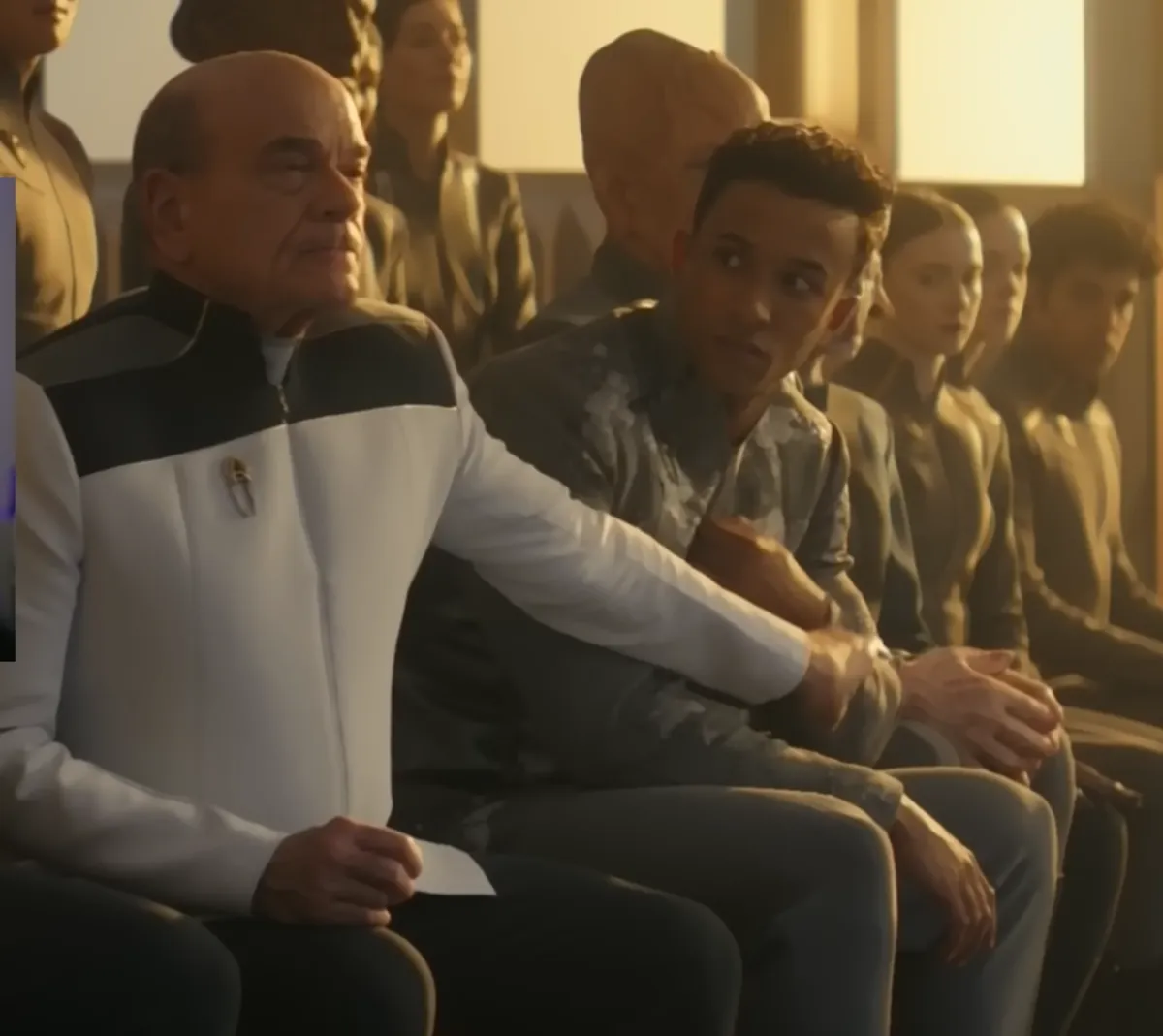 The Doctor, in a white Starfleet uniform, holds his arm across Caleb Mir's chest, feigning an attempt to restrain him, among a row of seated cadets.