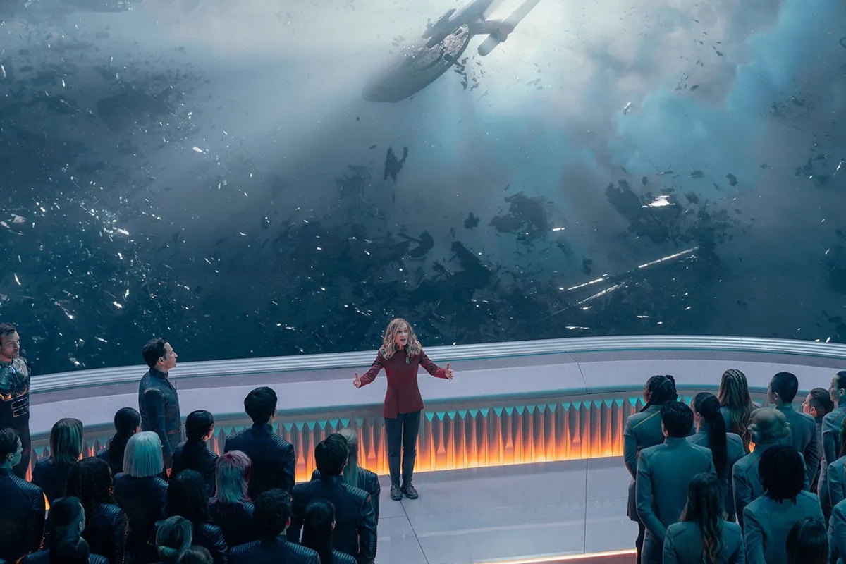 Chancellor Ake addresses a crowd of cadets on a balcony, a wrecked starship and debris drifting in the sky beyond.