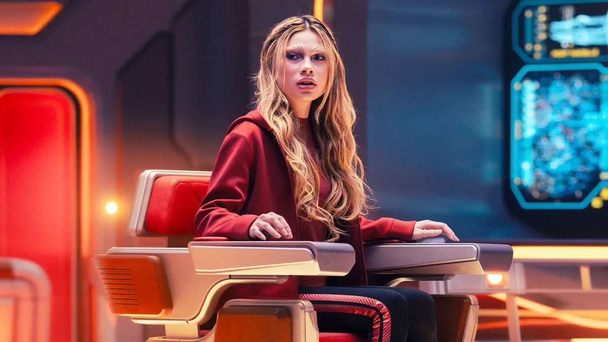 Lythe sitting in the captain's chair, in a red hooded jacket.