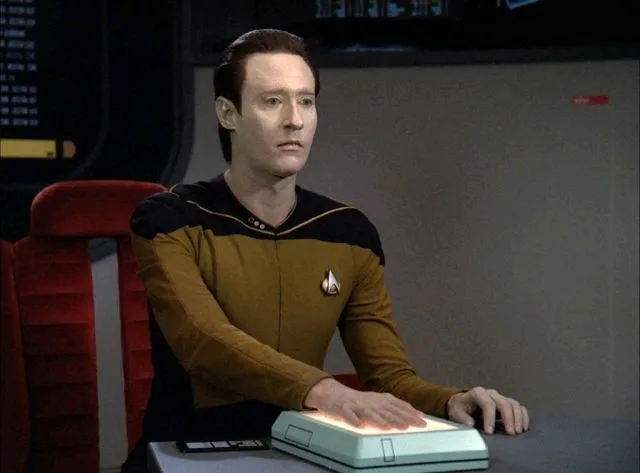Data seated at the hearing table, his hand resting on a panel, in "The Measure of a Man".